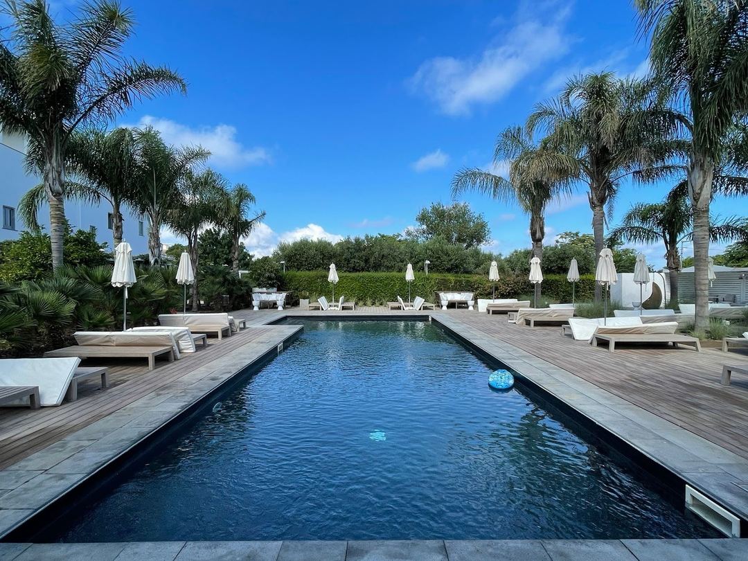 The hotel's rectangular swimming pool with wooden deck, sun loungers, white umbrellas, and palm trees under a clear blue sky.