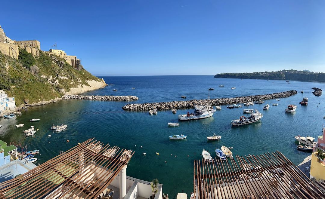 A panoramic coastal view of a Mediterranean harbor with moored fishing boats, rocky breakwaters, and hillside architecture under clear blue skies.
