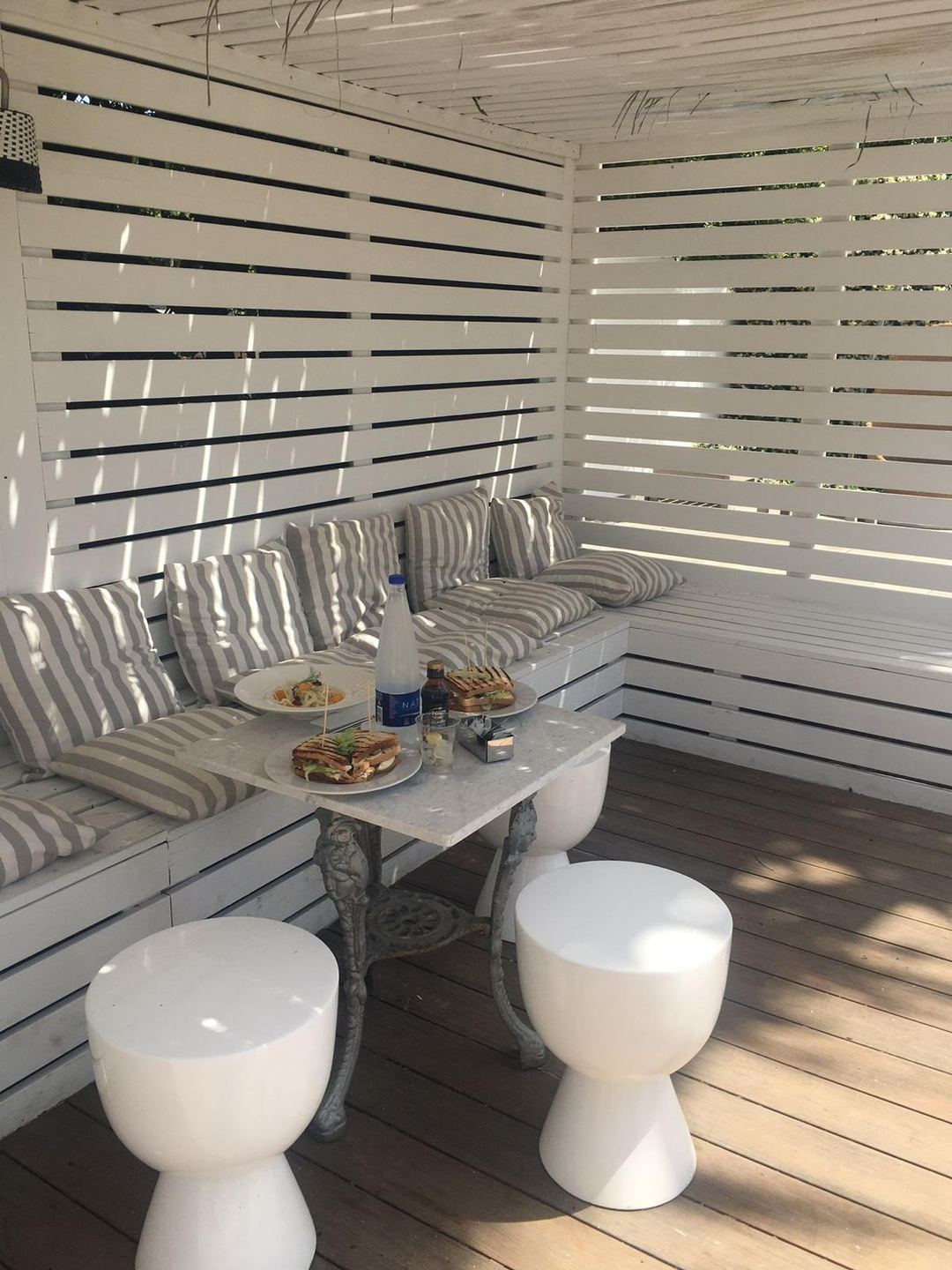 A modern outdoor lounge area with striped cushioned seating, white sculptural stools, and a marble table set with food and beverages, enclosed by horizontal slat screening.