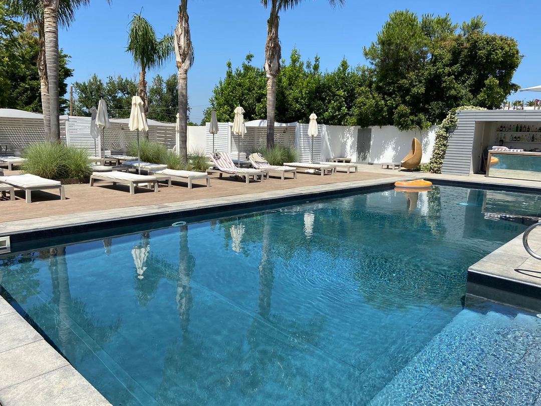 The hotel's outdoor swimming pool with wooden deck, white umbrellas, lounge chairs, and palm trees under clear blue sky.