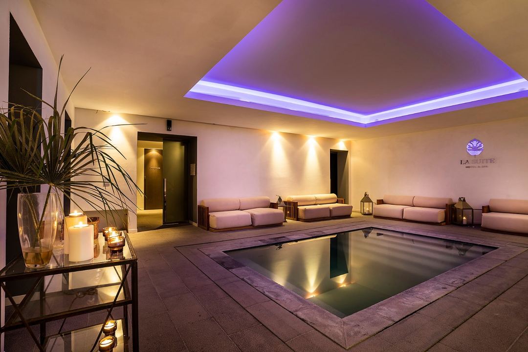 The spa's luxurious indoor pool area features ambient purple ceiling lighting, lounge seating, and decorative plants at La Suite Boutique Hotel & Spa.