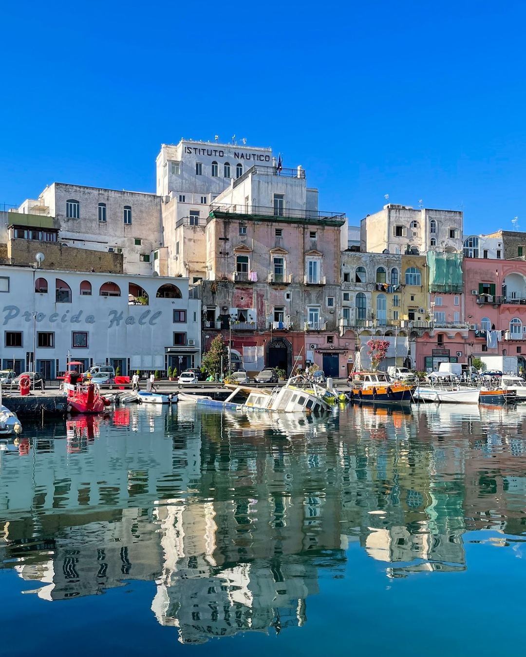 Colorful waterfront buildings and fishing boats reflected in calm harbor waters under clear blue sky.